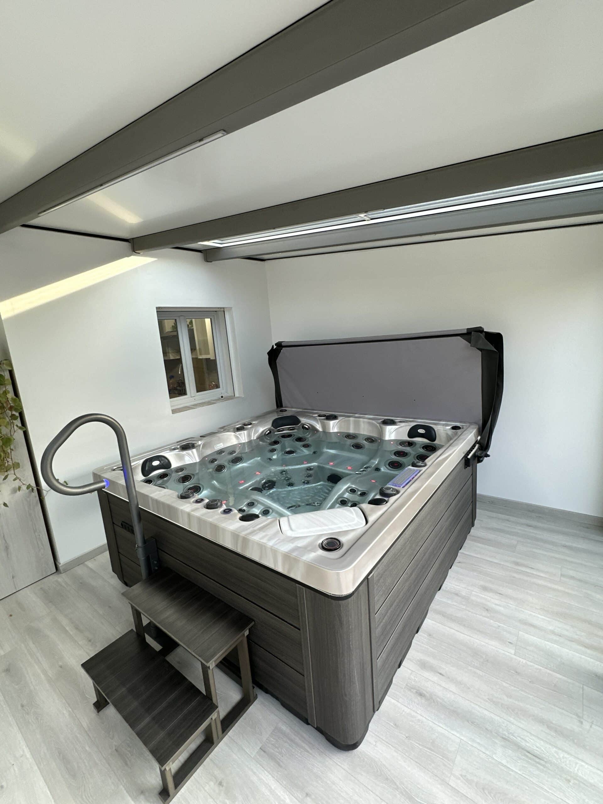 Jacuzzi intérieur moderne dans une pièce lumineuse. Le spa gris foncé est équipé de marches, d'une rampe éclairée et d'un couvercle relevé.