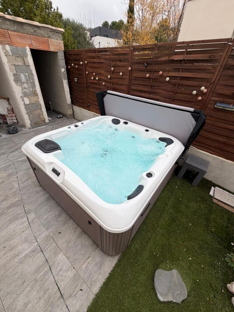 Grand spa jacuzzi extérieur bouillonnant (eau turquoise) sur une terrasse. Cabine marron, couverture grise. Clôture en bois et gazon synthétique.