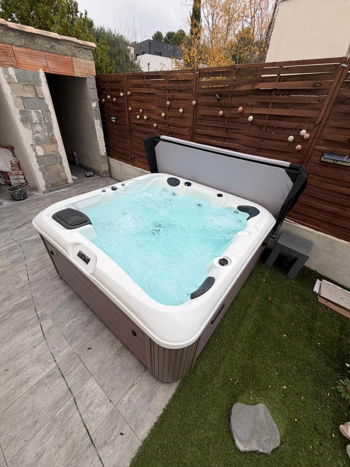 Grand spa jacuzzi extérieur bouillonnant (eau turquoise) sur une terrasse. Cabine marron, couverture grise. Clôture en bois et gazon synthétique.