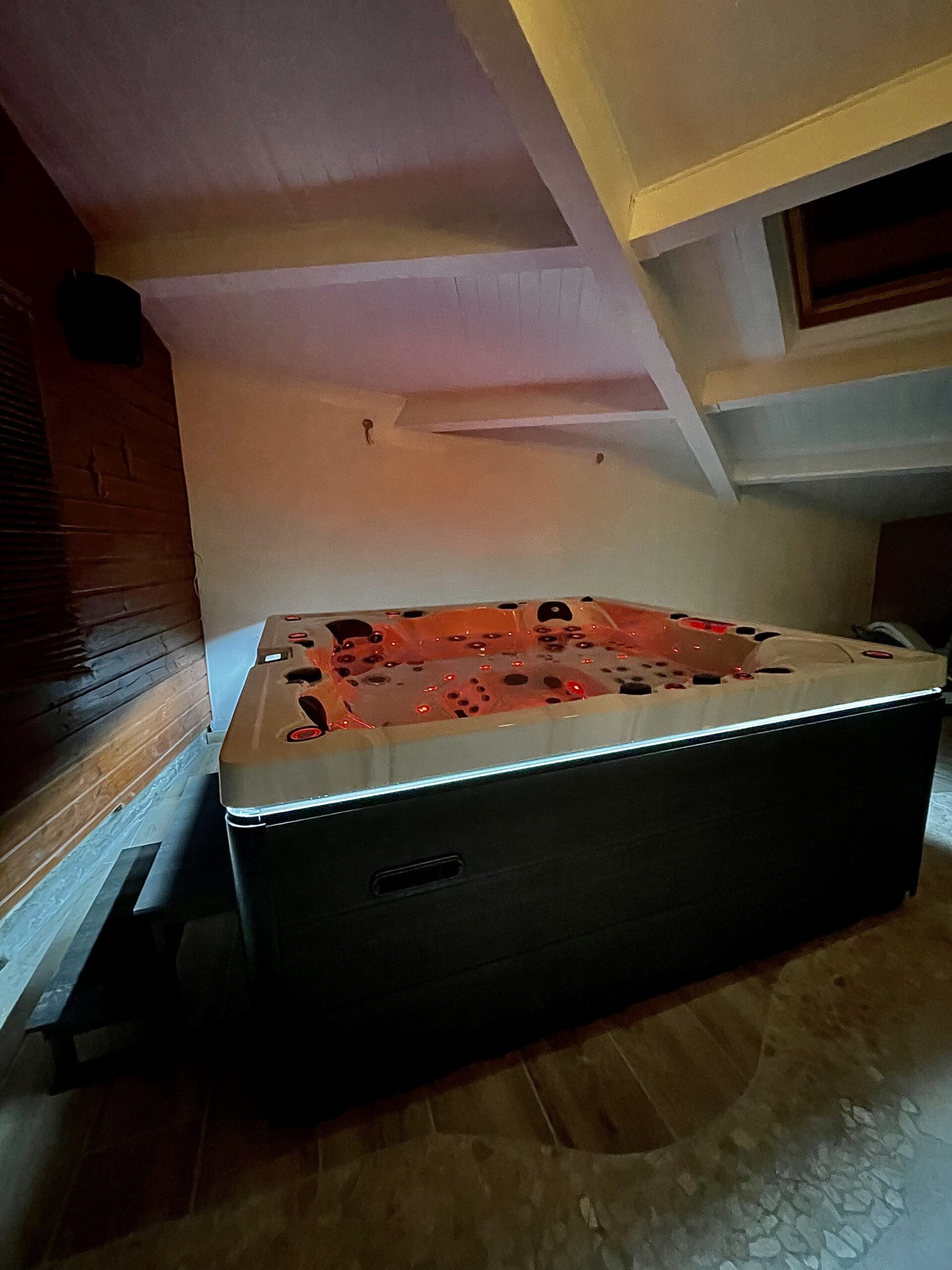 Luxueux jacuzzi intérieur sous un plafond mansardé. L'eau rougeoyante et l'éclairage LED bleu créent une ambiance romantique dans cet espace bois.
