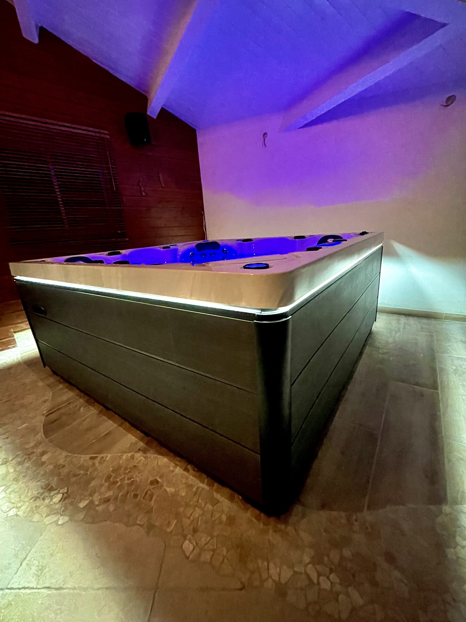 Jacuzzi moderne intérieur. Éclairage LED bleu du spa, coque claire, jupes sombres, sous un plafond incliné baigné de lumière violette.