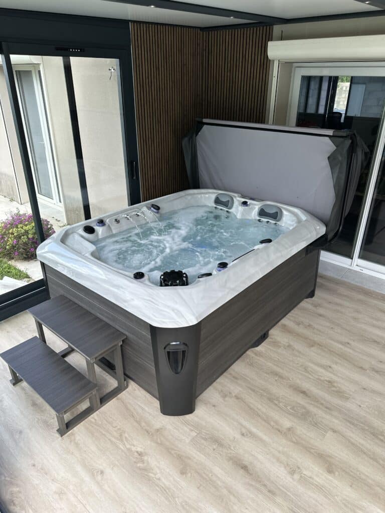 Grand spa jacuzzi contemporain installé en intérieur, entouré de baies vitrées. Habillage gris bois, murs à lattes et marches d'accès.
