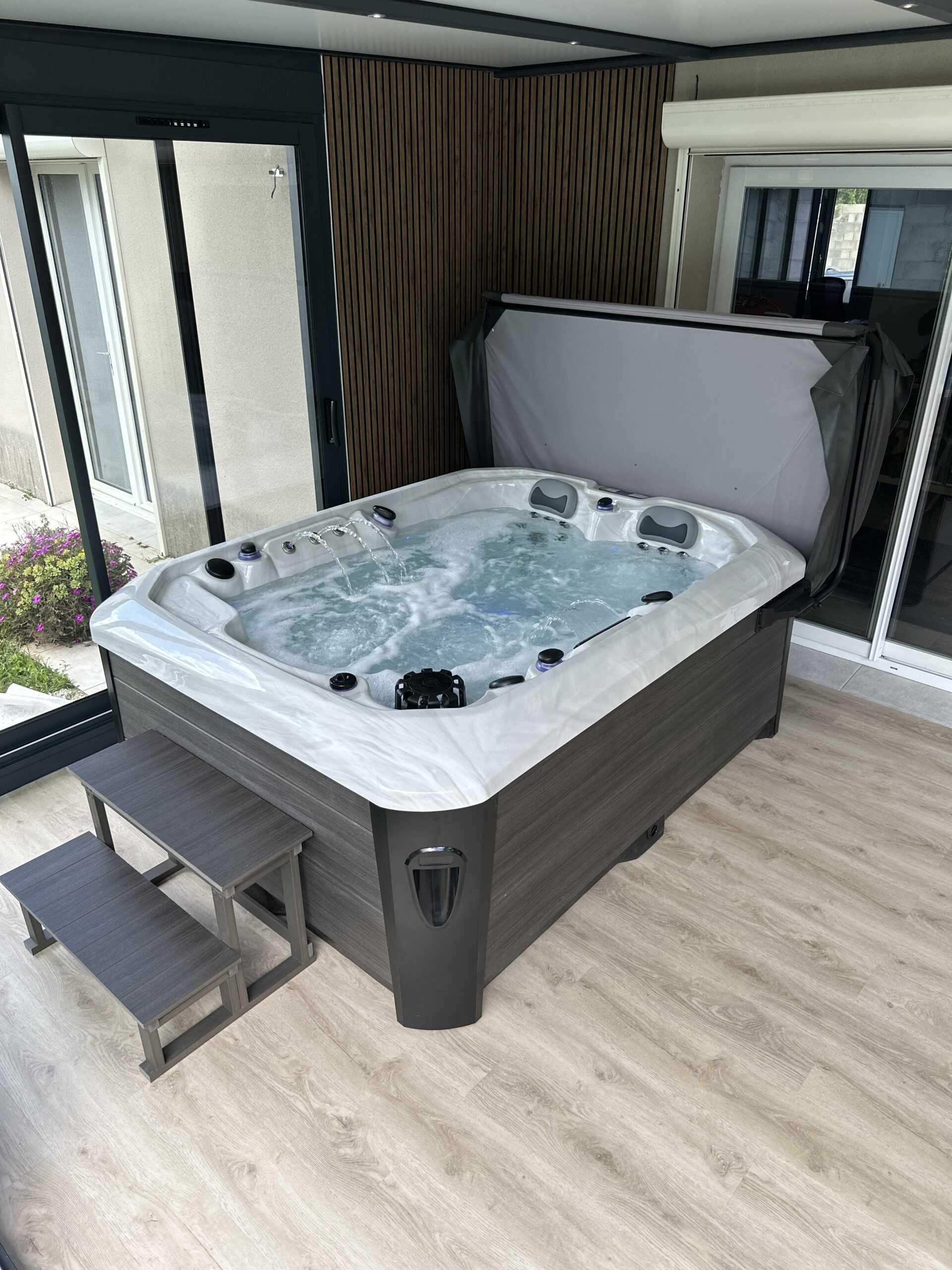 Grand spa jacuzzi contemporain installé en intérieur, entouré de baies vitrées. Habillage gris bois, murs à lattes et marches d'accès.