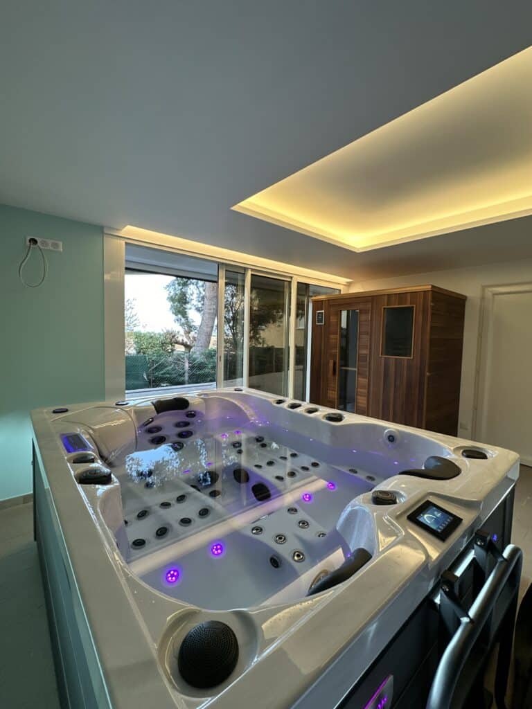 Spa Jacuzzi luxe intérieur et Sauna : L'espace bien-être moderne Spa jacuzzi moderne blanc illuminé par LED violettes. Salle de bien-être luxe avec sauna en bois, plafond lumineux et vue sur jardin.
