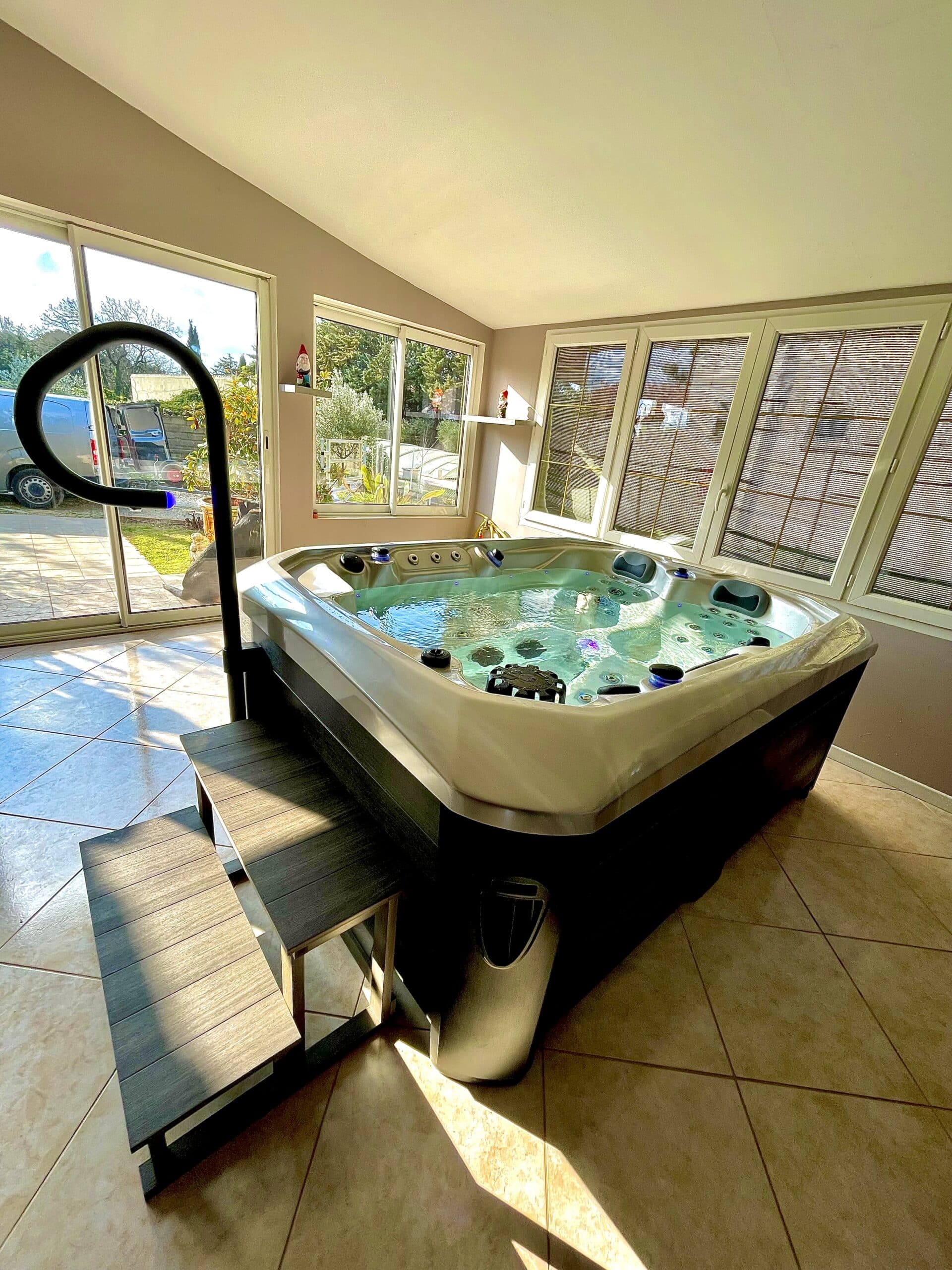 Jacuzzi intérieur moderne et ses marches en composite, éclairé par la lumière du soleil entrant dans la véranda vitrée. Ambiance détente.