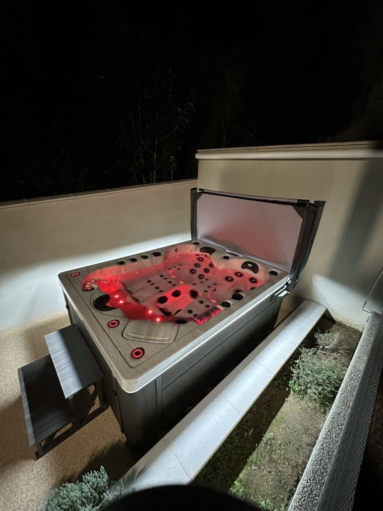 Spa de Nuit : Jacuzzi Extérieur à Éclairage Rouge Spa de nuit, cuve illuminée en rouge vif. Le jacuzzi extérieur possède des marches d'accès et une couverture relevée pour la détente.