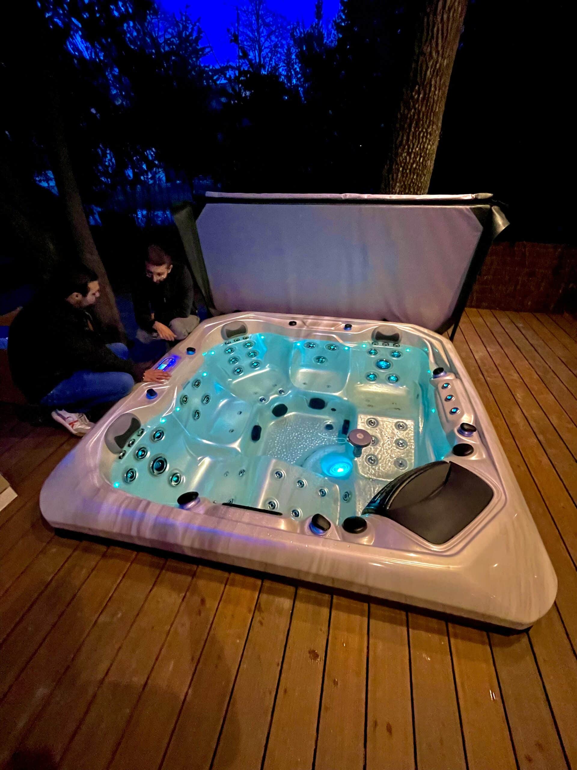 Bain à remous extérieur sur terrasse en bois, illuminé par des LED turquoise. Deux hommes préparent le spa la nuit tombée.