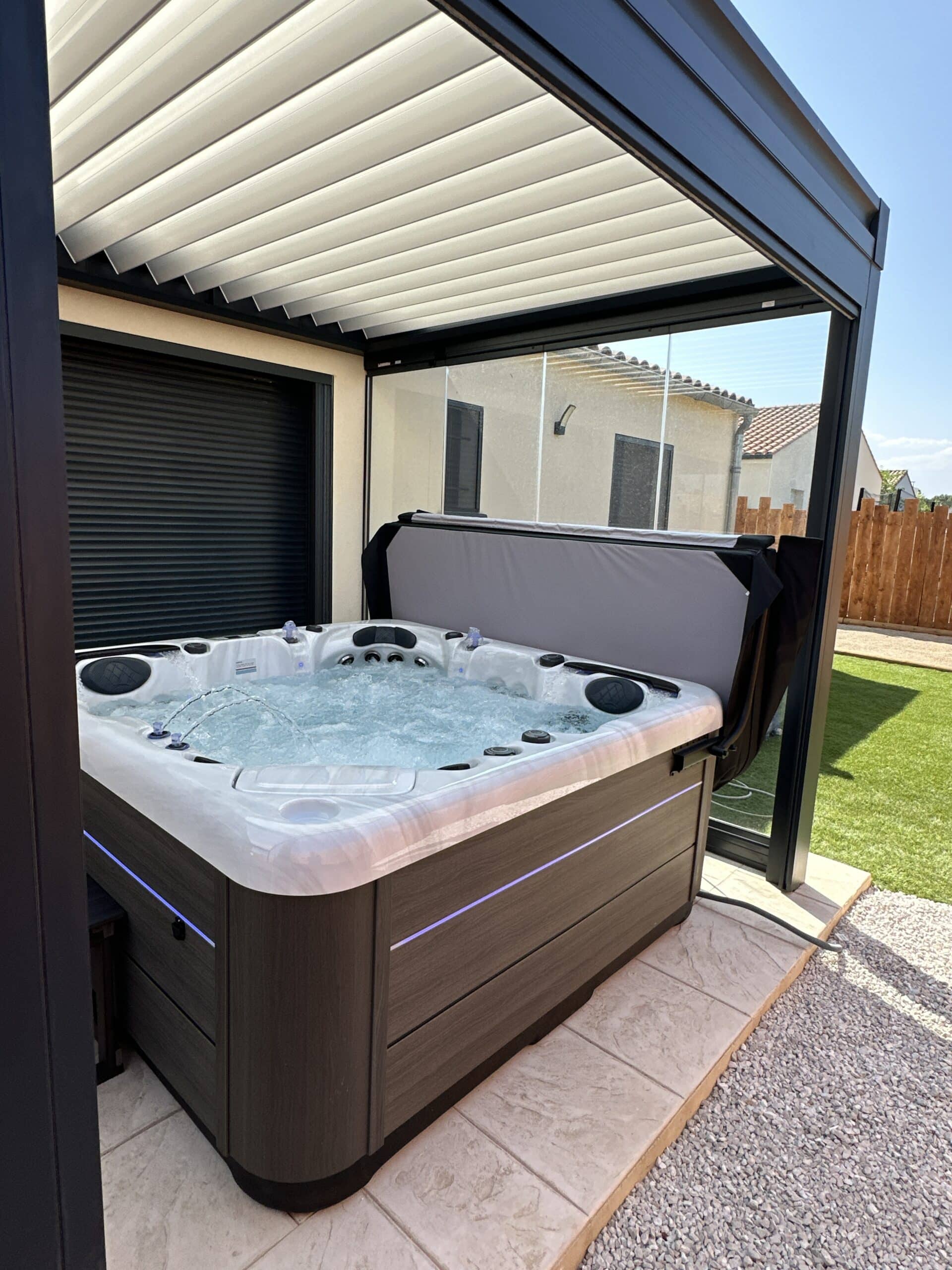 Spa moderne avec éclairage bleu sous une pergola bioclimatique en aluminium noir. Lames orientables et parois vitrées sur une terrasse.