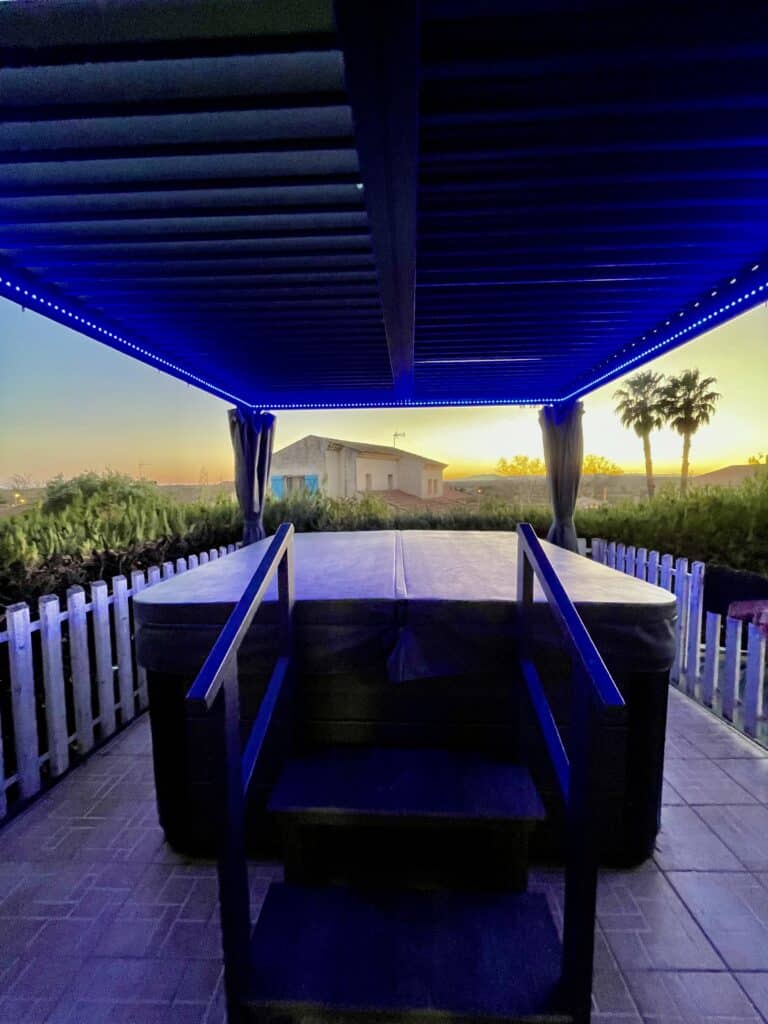 Spa extérieur de luxe : Détente sous les LED bleues au coucher du soleil Jacuzzi extérieur couvert sous une pergola sombre éclairée par des LED bleues vives. Vue symétrique sur le coucher de soleil et deux palmiers.