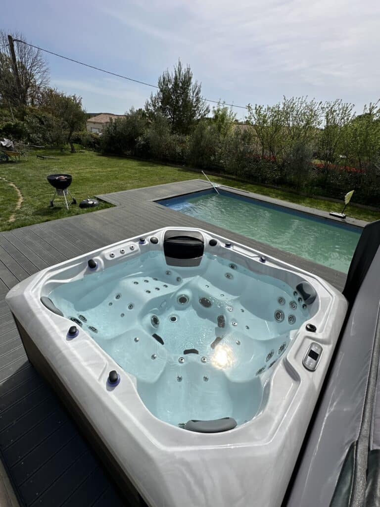 Grand jacuzzi moderne sur terrasse composite gris foncé, avec piscine attenante et jardin verdoyant ensoleillé. BBQ visible.