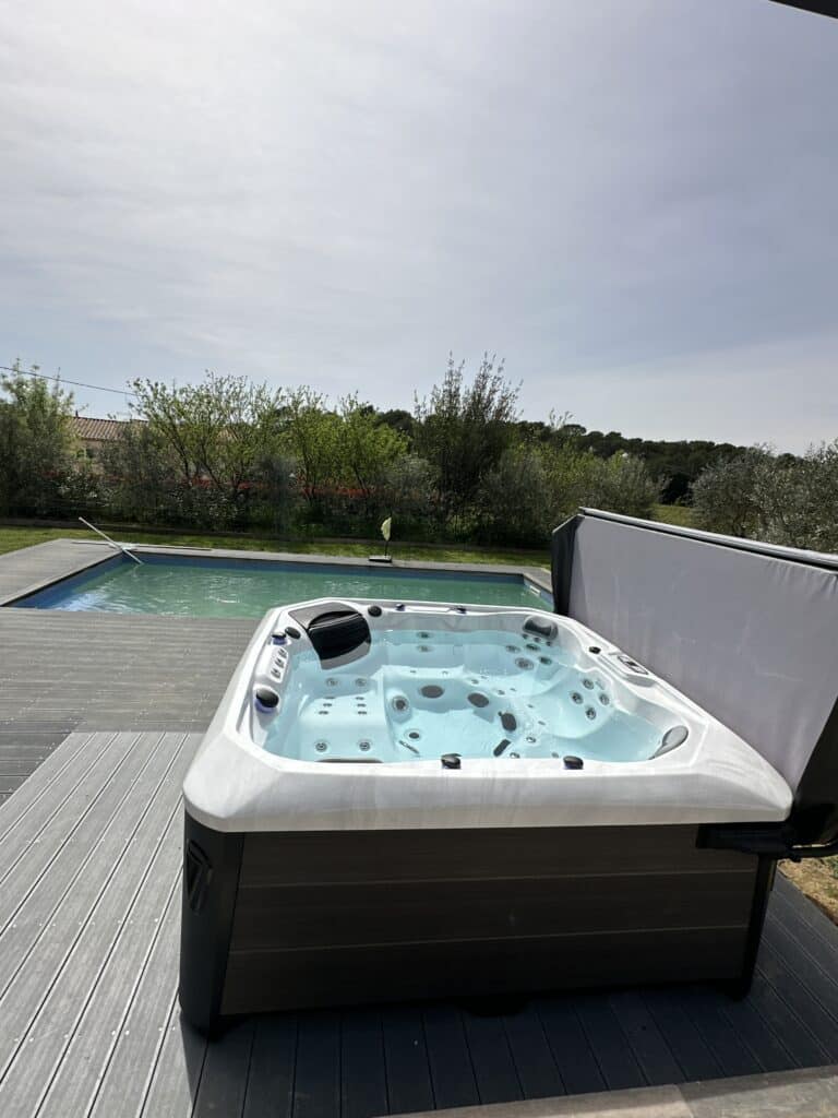 Spa jacuzzi et piscine sur terrasse composite moderne Jacuzzi blanc moderne sur une terrasse composite grise. Le spa est face à une piscine et un jardin verdoyant, sous un ciel lumineux.