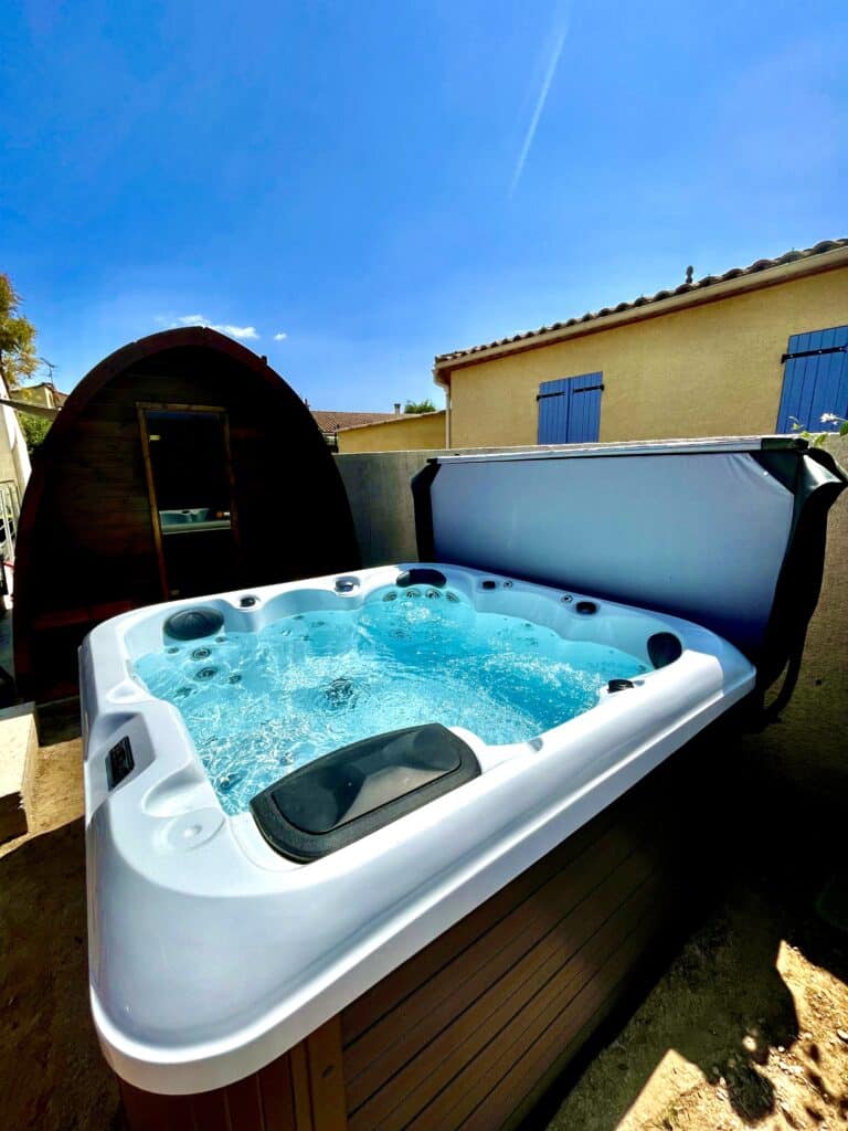 Spa jacuzzi et sauna extérieur sous un ciel bleu d'été Luxueux spa jacuzzi bouillonnant près d'un sauna tonneau en bois, dans un jardin baigné de soleil. Maison ocre et ciel bleu.