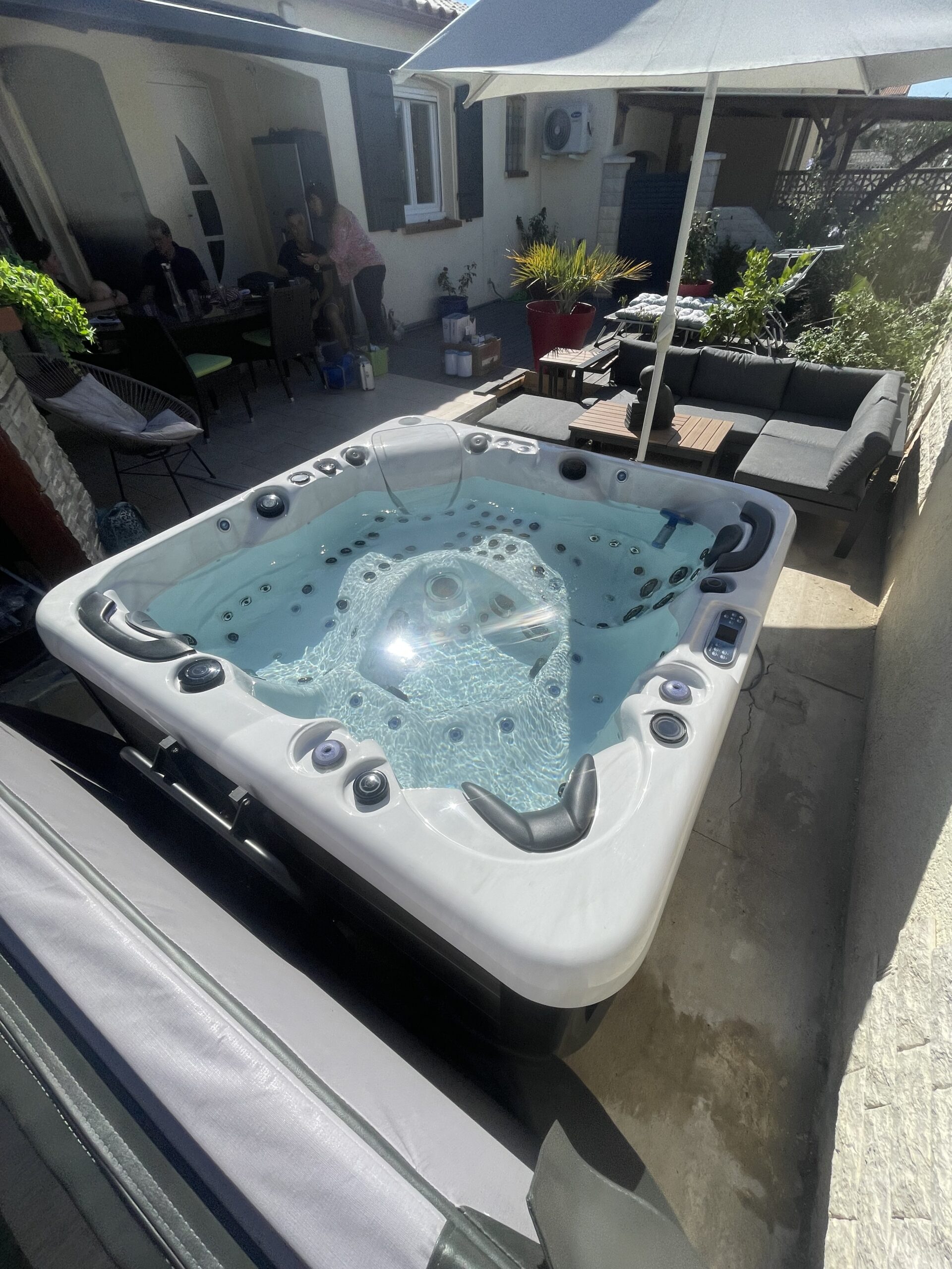 Grand Jacuzzi extérieur blanc rempli d'eau pétillante, installé sur une terrasse ensoleillée. Un salon de jardin et des convives sont à l'ombre.