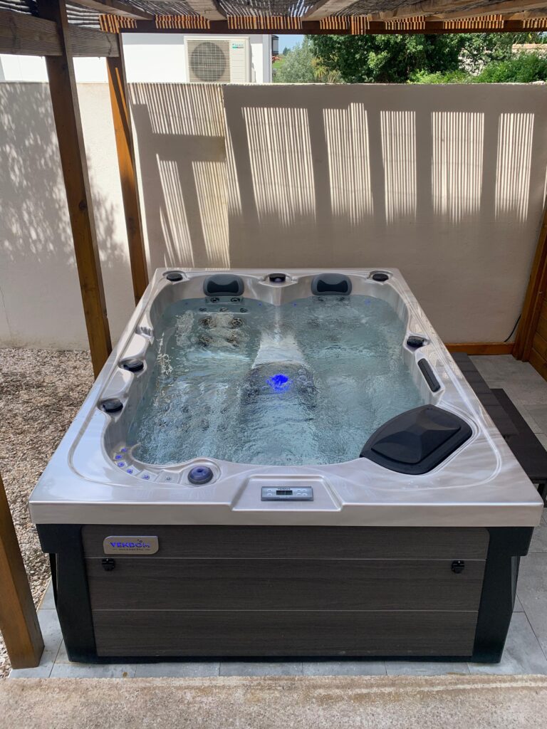 Jacuzzi VENDOM moderne sur terrasse, eau bouillonnante avec LED bleue. L'éclairage naturel crée des ombres de pergola sur le mur blanc.