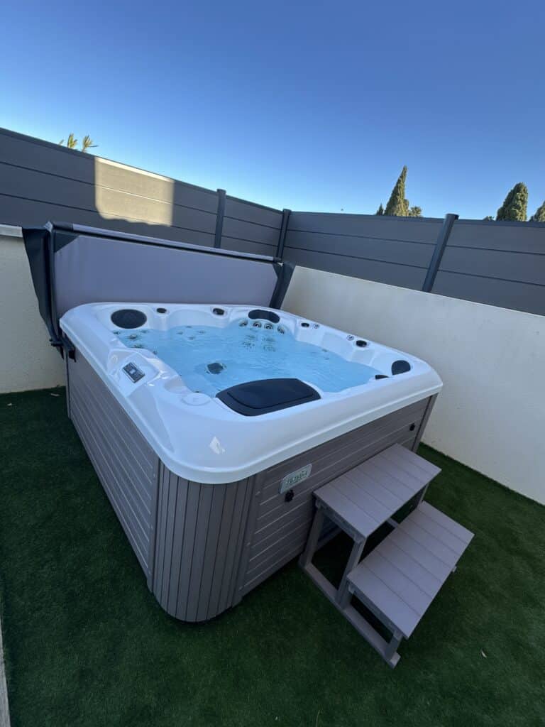 Spa extérieur Vendom moderne : installation sur terrasse gazonnée. Spa Vendom (Marque Egea) en extérieur avec marches grises, couverture ouverte, sur gazon synthétique contre une clôture anthracite.