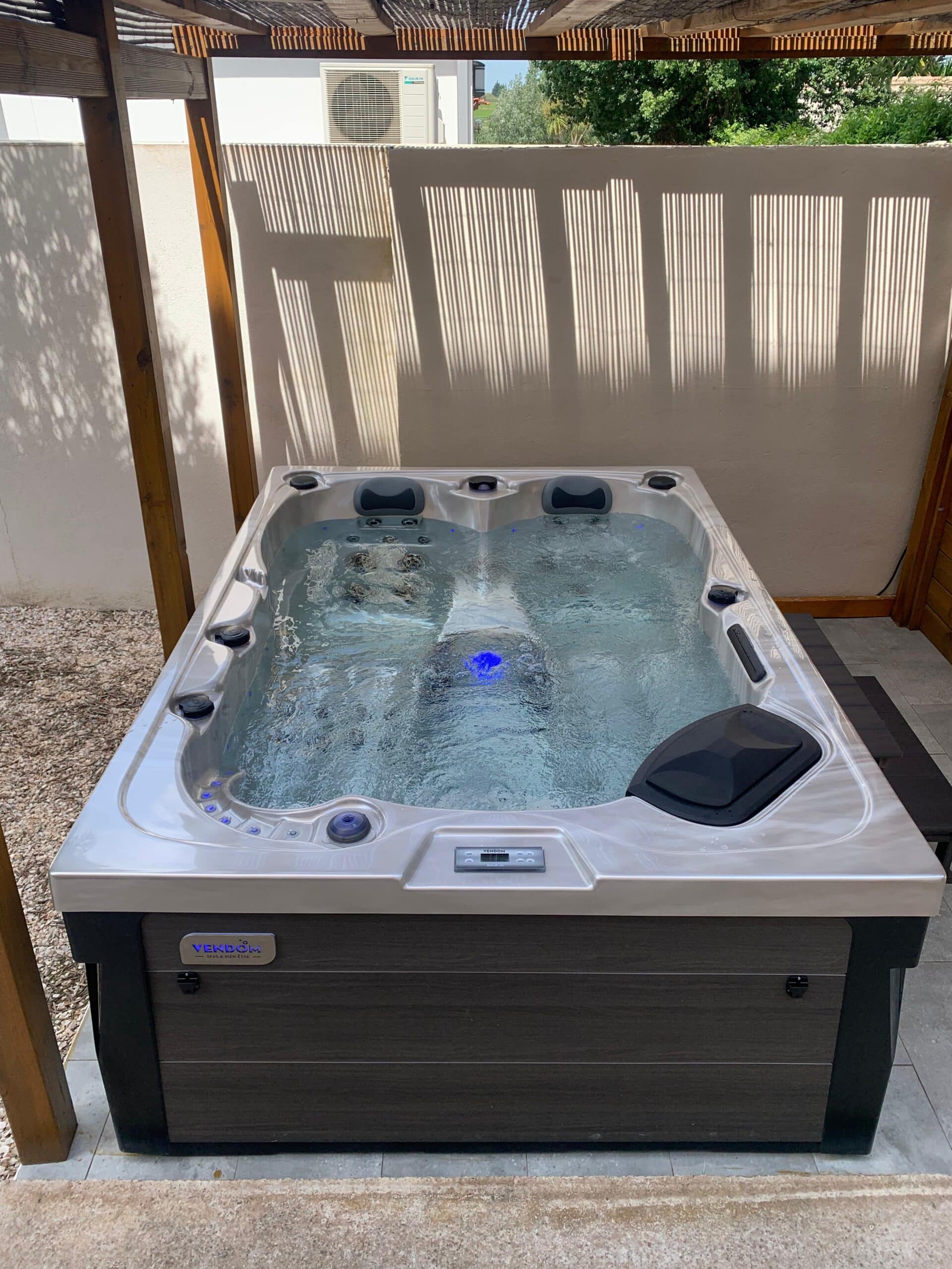 Jacuzzi VENDOM moderne sur terrasse, eau bouillonnante avec LED bleue. L'éclairage naturel crée des ombres de pergola sur le mur blanc.