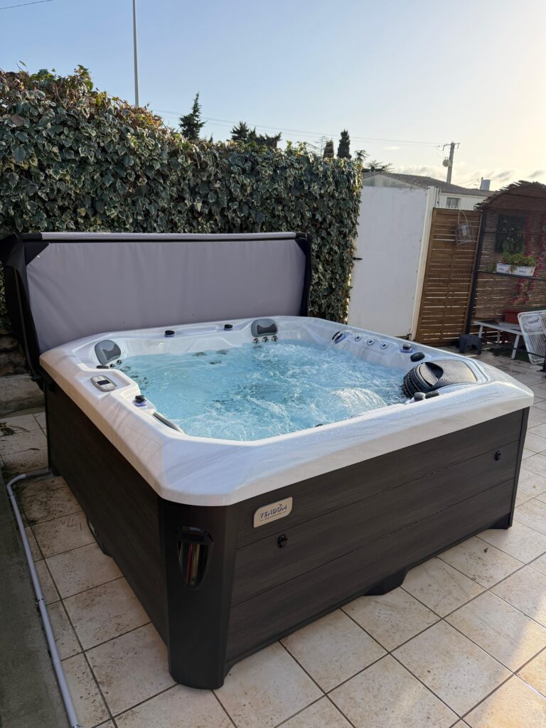 Grand spa Vendôm extérieur en fonctionnement, avec eau bouillonnante sur une terrasse carrelée. Le couvercle relevé offre une intimité contre la haie de lierre.