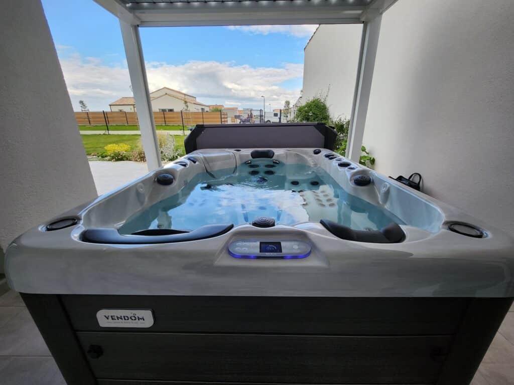 Spa VENDOM extérieur pour terrasse | Détente et bien-être. Spa VENDOM moderne (19.5°C) sur une terrasse extérieure abritée. La cuve grise est remplie d'eau, offrant une vue dégagée sur le ciel bleu.