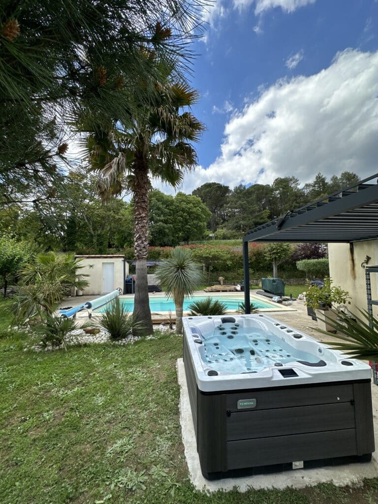 Spa Jacuzzi Vendôme : Piscine et Jardin Méditerranéen. Spa Vendôme noir et gris rempli, installé sur terrasse. Vue sur une piscine, des palmiers et un jardin luxuriant sous ciel bleu.