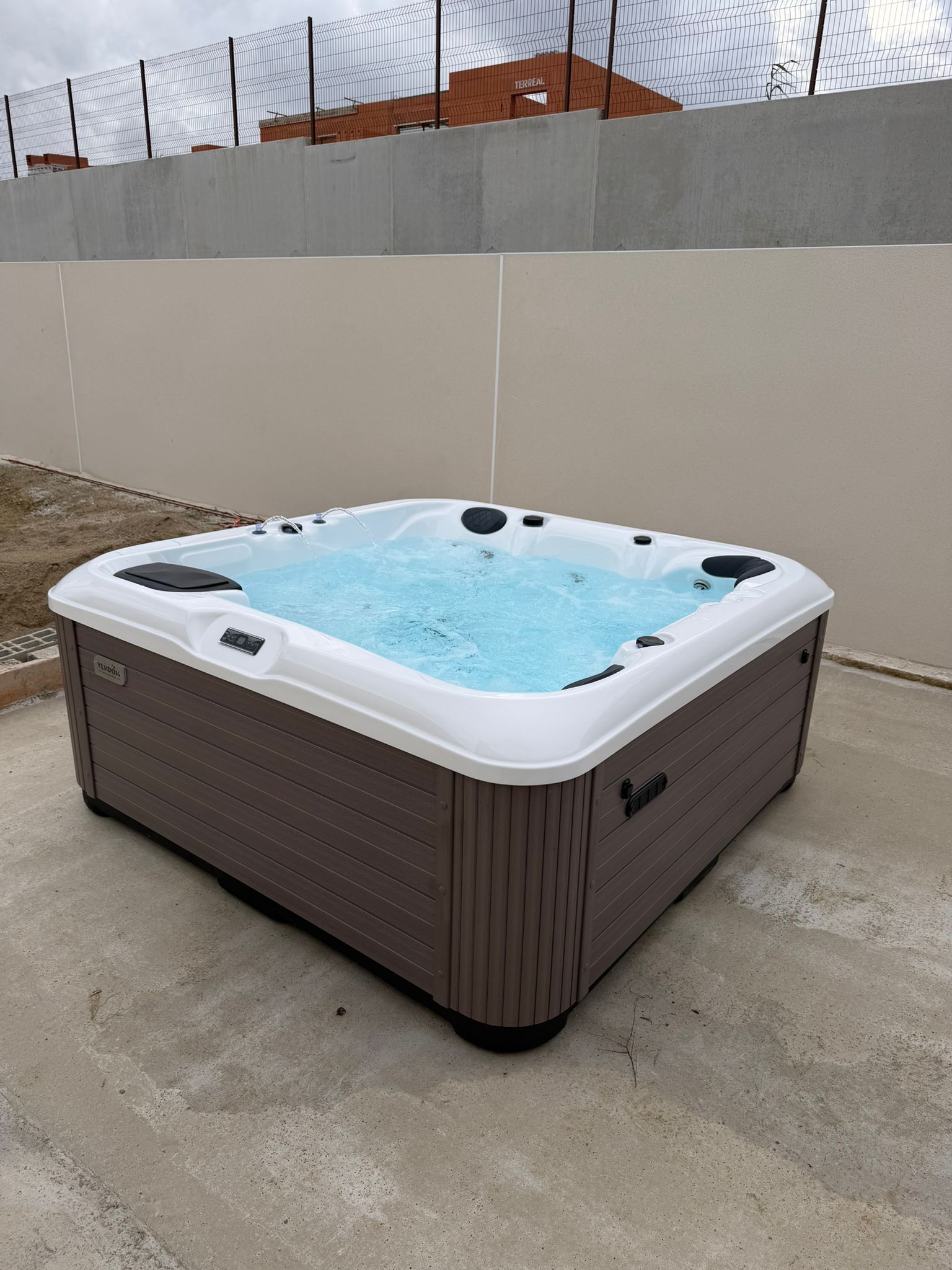 Spa Ver dok rectangulaire, coque blanche et panneau marron effet bois, installé sur dalle de béton. L'eau bleue bouillonne.