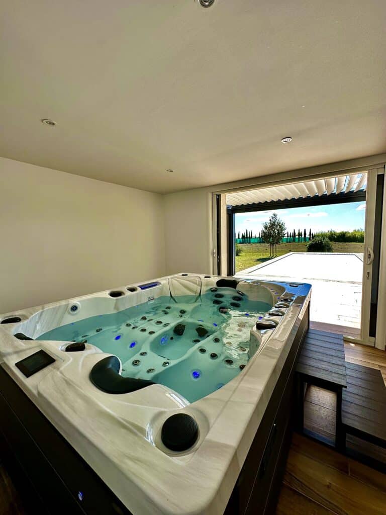 Spa de nage intérieur luxe et jacuzzi moderne avec vue. Spa de nage intérieur moderne avec jets et lumières LED bleues. La pièce s'ouvre sur une grande terrasse en carrelage et un paysage ensoleillé.
