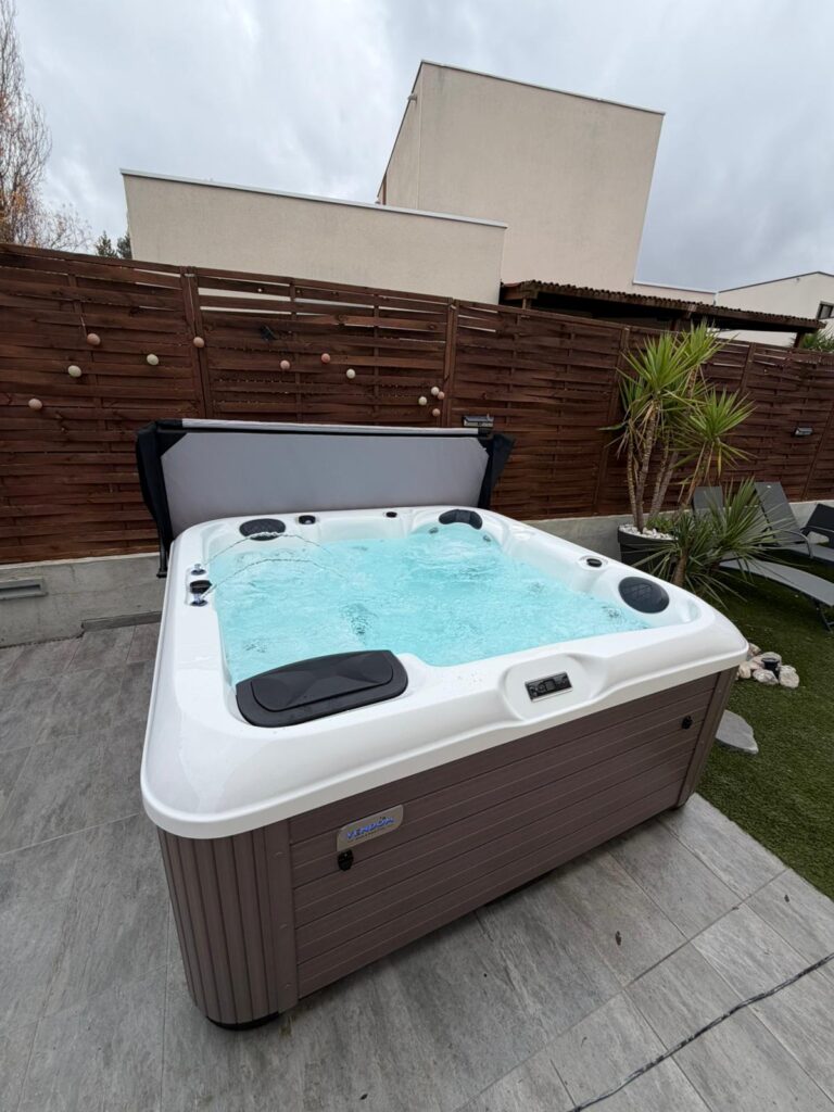 Spa extérieur Vendôm : luxe et détente sur terrasse. Spa Vendôm extérieur marron sur terrasse carrelée. Le bain à remous est actif, la couverture relevée, clôture bois foncé en arrière-plan.