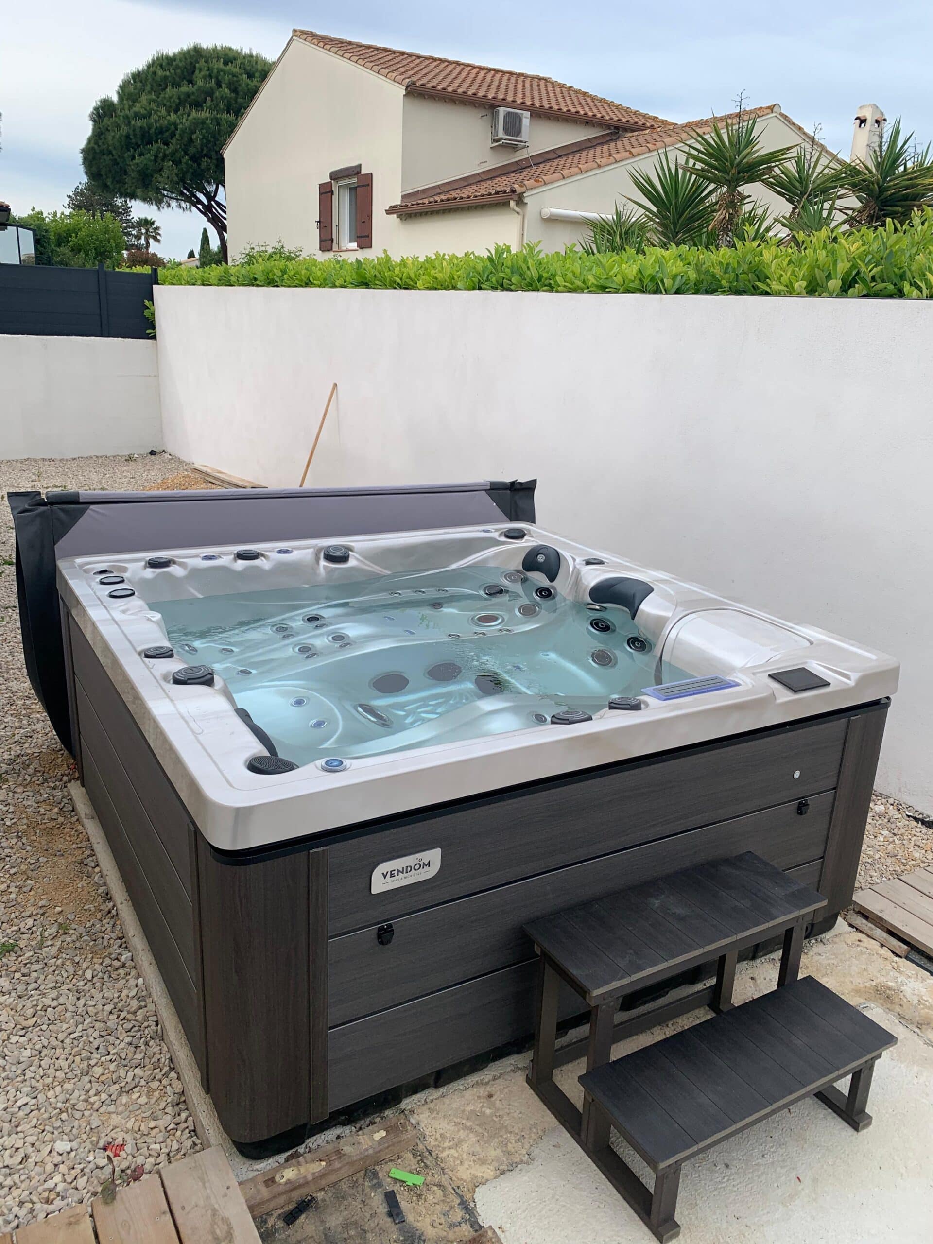 Spa extérieur Vendom, moderne et gris anthracite, installé sur une terrasse en gravier près d'un mur blanc et d'une maison.