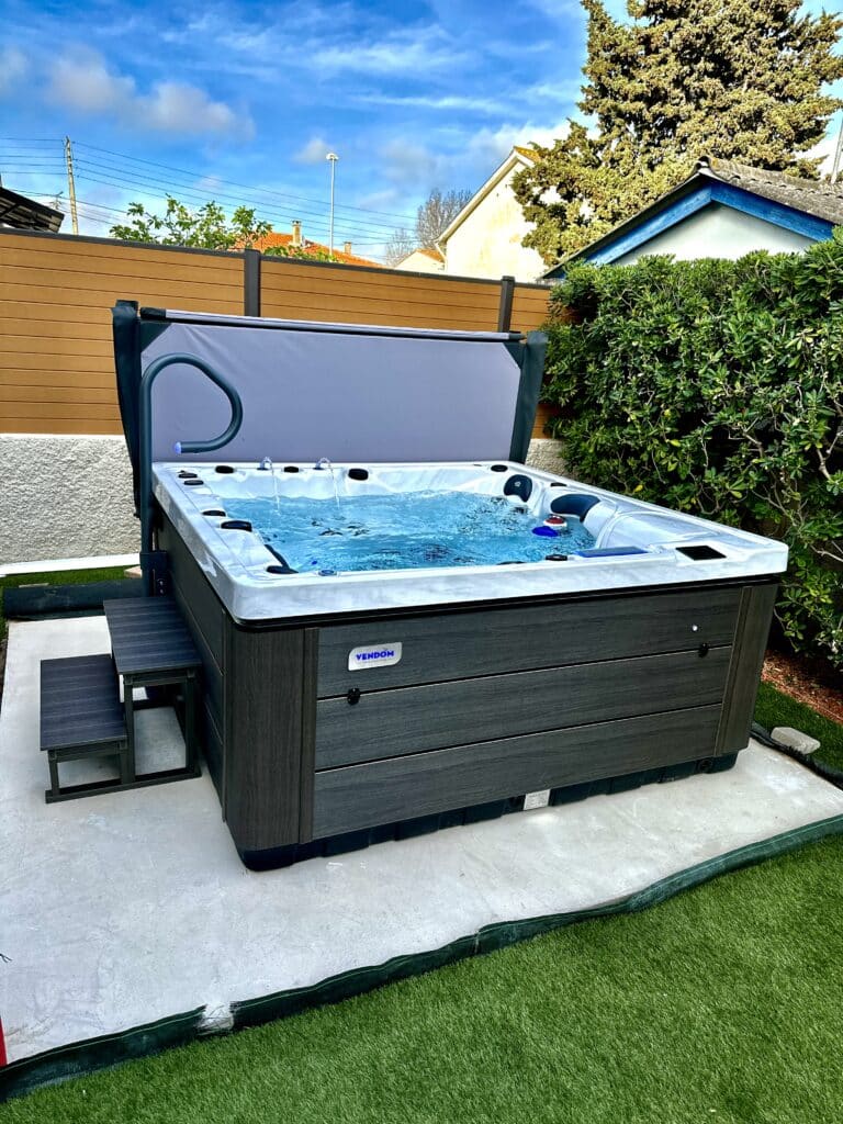 Spa Vendom : Détente et bien-être en plein air. Spa Vendom de luxe en extérieur, jets d'eau, revêtement gris foncé, marches, main courante et couverture relevée. VENDOM SPAS & BIEN ÊTRE.